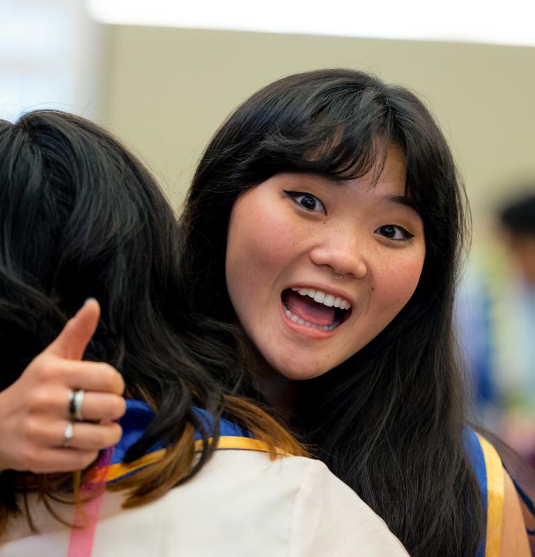 A student gives a thumbs-up while hugging a friend at a campus event and smiling enthusiastically toward the camera.