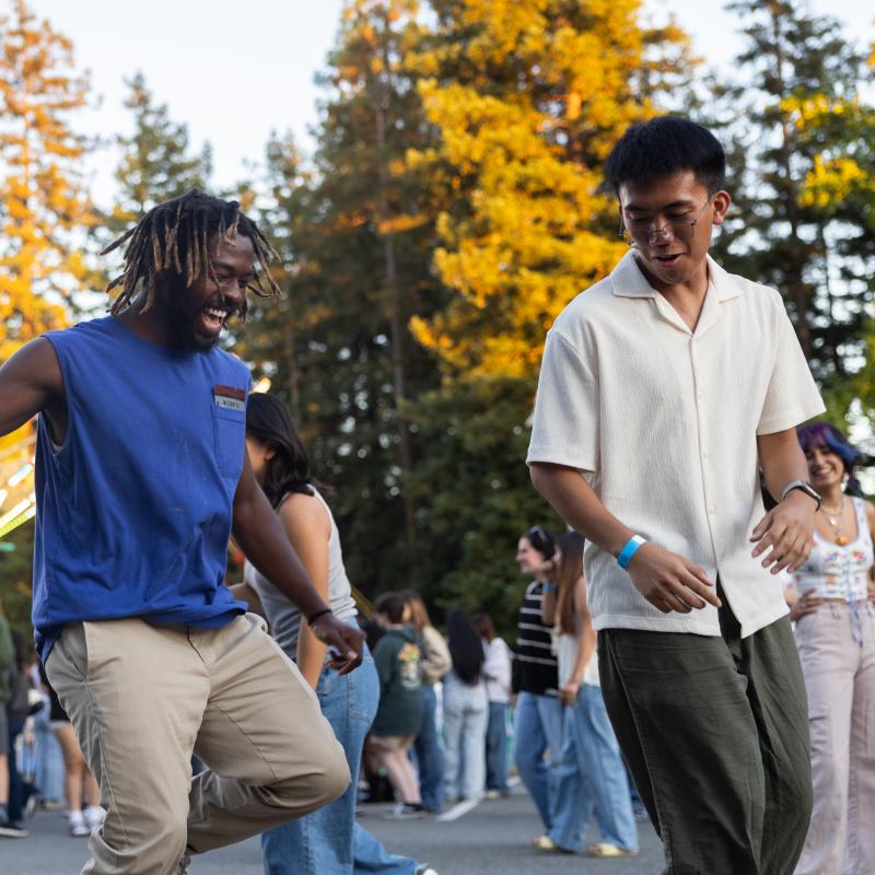 Students dance and enjoy live entertainment during an outdoor campus celebration surrounded by classmates and trees.